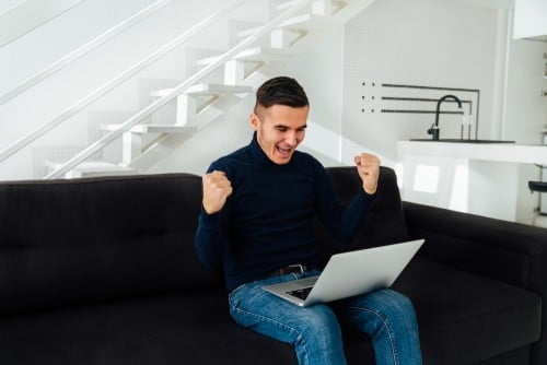 jeune homme heureux de ses gains assis sur un sofa avec son ordinateur portable sur les genoux